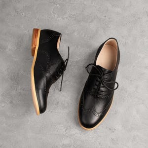 K&ouml;nnte beinhalten: Ein Paar schwarze Lederschuhe im Oxford-Stil mit klassischem Brogue-Muster, abgerundeter Zehenpartie, schwarzer Schn&uuml;rung sowie einem hellbraunen Holzabsatz und einer hellen Sohle. Die Schuhe sind in verschiedenen Winkeln angeordnet.