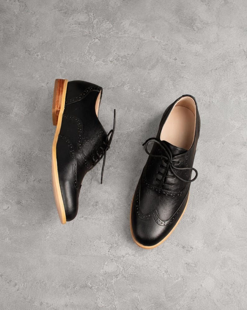 K&ouml;nnte beinhalten: Ein Paar schwarze Lederschuhe im Oxford-Stil mit klassischem Brogue-Muster, abgerundeter Zehenpartie, schwarzer Schn&uuml;rung sowie einem hellbraunen Holzabsatz und einer hellen Sohle. Die Schuhe sind in verschiedenen Winkeln angeordnet.