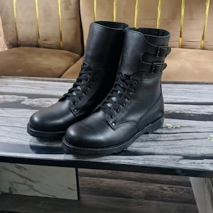 Puede incluir: Un par de botas negras de media caña con punta redondeada, cordones negros y dos correas con hebilla ajustables en la parte superior de la caña. Las botas tienen un acabado liso, una suela resistente y un diseño que llega sobre el tobillo.