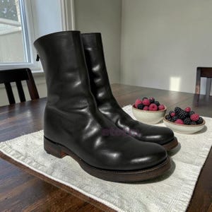 May include: A pair of black leather mid-calf boots featuring a smooth finish, a rounded toe, and a dark brown stacked heel. The boots have a vertical zipper closure on the back of the shaft and a sturdy sole, presented with two small bowls of berries.