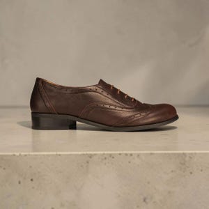 K&ouml;nnte beinhalten: Ein dunkelbrauner Oxford-Schuh aus Leder mit abgerundeter Schuhspitze, niedrigem Blockabsatz und dekorativen Brogue-Perforationen entlang der N&auml;hte. Der Schuh ist mit d&uuml;nnen, hellbraunen Schn&uuml;rsenkeln durch &Ouml;sen am Vorderblatt versehen.