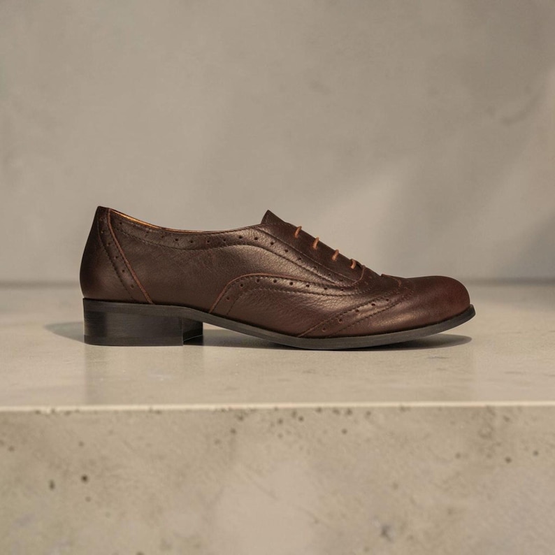K&ouml;nnte beinhalten: Ein dunkelbrauner Oxford-Schuh aus Leder mit abgerundeter Schuhspitze, niedrigem Blockabsatz und dekorativen Brogue-Perforationen entlang der N&auml;hte. Der Schuh ist mit d&uuml;nnen, hellbraunen Schn&uuml;rsenkeln durch &Ouml;sen am Vorderblatt versehen.