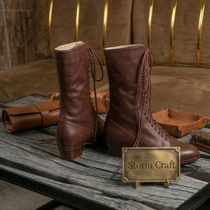 May include: A pair of brown leather lace-up boots with a mid-calf height and a low block heel. A small rectangular brass plaque stands in front of the boots, featuring the engraved text Storm Craft in a serif font, with matching leather accessories.
