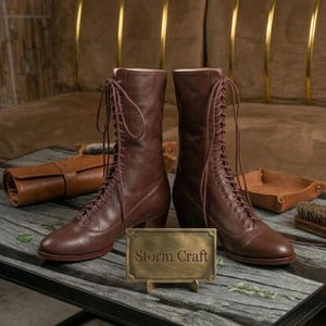 May include: A pair of mid-calf brown leather lace-up boots with rounded toes and stacked heels. The boots feature long matching laces tied at the top. A rectangular brass plaque stands in front of the boots with the text Storm Craft engraved.