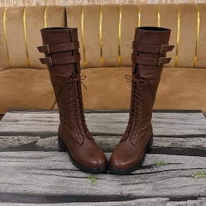 May include: A pair of knee-high brown leather boots featuring a lace-up front design with thin brown laces. Each boot includes two adjustable straps with metal buckles positioned at the top shaft and a thick black sole with a low heel.