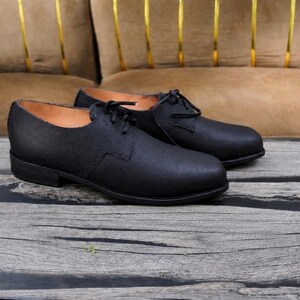 K&ouml;nnte beinhalten: Ein Paar schwarze Schn&uuml;rschuhe mit matter Oberfl&auml;che, abgerundeter Schuhspitze, flachem Absatz und beigefarbenem Innenfutter. Die Schuhe haben ein schlichtes, klares Design mit sichtbaren N&auml;hten an den Seitenteilen und schwarzen Schn&uuml;rsenkeln.
