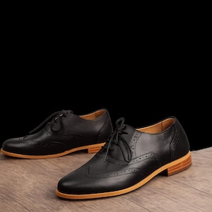 K&ouml;nnte beinhalten: Ein Paar schwarze Oxford-Schuhe aus Leder mit klassischem Wingtip-Brogue-Muster, d&uuml;nnen schwarzen Schn&uuml;rsenkeln und einer markanten hellbraunen Gummisohle. Die Schuhe haben eine abgerundete Spitze, einen flachen Absatz und dekorative Perforationen.