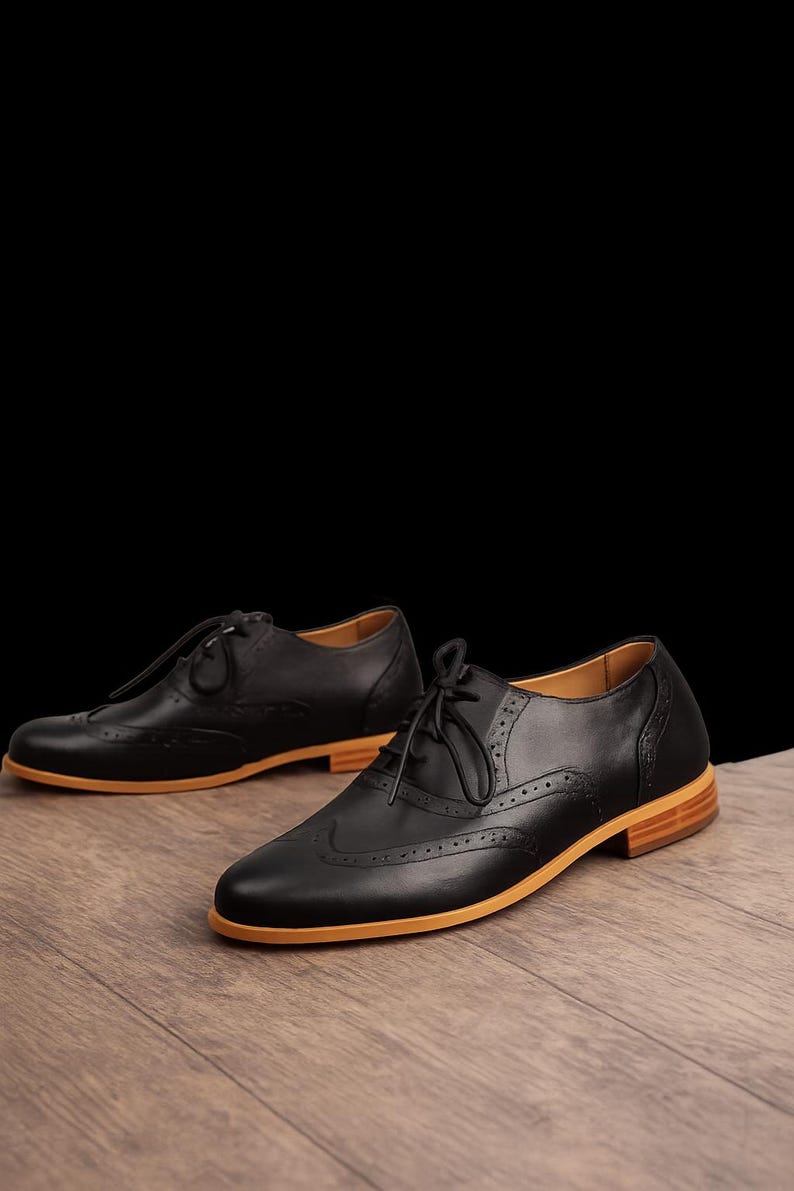 K&ouml;nnte beinhalten: Ein Paar schwarze Oxford-Schuhe aus Leder mit klassischem Wingtip-Brogue-Muster, d&uuml;nnen schwarzen Schn&uuml;rsenkeln und einer markanten hellbraunen Gummisohle. Die Schuhe haben eine abgerundete Spitze, einen flachen Absatz und dekorative Perforationen.
