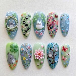 May include: A set of ten press-on nails featuring intricate 3D art inspired by Studio Ghibli characters. Designs include Totoro figures, soot sprites, lotus flowers, butterflies, and floral patterns in shades of green, blue, pink, and white.