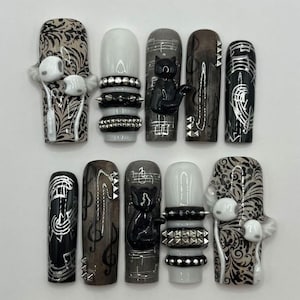 May include: A set of ten press-on nails featuring a black, white, and gray color palette. Designs include 3D white earbuds, silver studded bands, black cat silhouettes, and silver musical notes on black and gray backgrounds.