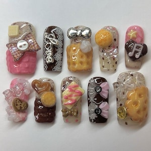May include: A set of ten press-on nails featuring 3D embellishments including miniature waffles, bows, hearts, buttons, and musical notes. The designs incorporate brown, pink, and cream color palettes with lace patterns, polka dots, and text.