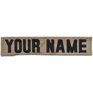 May include: A rectangular name patch featuring a camouflage pattern in shades of tan, brown, and green. The text "YOUR NAME" is embroidered in bold, black, capital letters across the center of the patch, which has a stitched border.