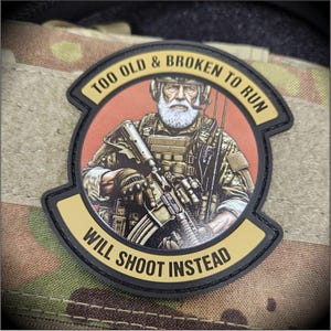 May include: A circular morale patch featuring a bearded individual in tactical gear holding a rifle. The text 'TOO OLD & BROKEN TO RUN' is arched at the top, and 'WILL SHOOT INSTEAD' is arched at the bottom, both in black on a tan background.