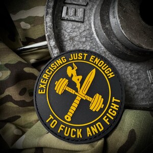 May include: A circular black patch with yellow text and graphics. The text reads EXERCISING JUST ENOUGH TO FUCK AND FIGHT. The graphic features a silhouette of a person climbing a barbell crossed with a sword, all in a bold yellow color scheme.