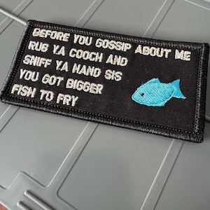 May include: A rectangular black embroidered patch with a stitched border. The text reads: BEFORE YOU GOSSIP ABOUT ME RUB YA COOCH AND SNIFF YA HAND SIS YOU GOT BIGGER FISH TO FRY. A small, light blue embroidered fish icon is positioned on the right.