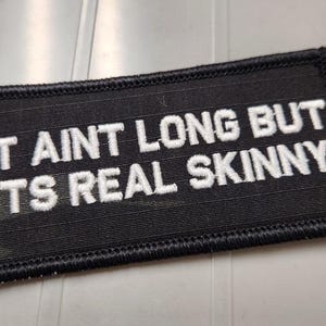 May include: A rectangular black embroidered patch with a stitched border. The patch features white text in a bold, sans-serif font that reads: IT AINT LONG BUT ITS REAL SKINNY. The design is centered on the fabric with a clean, uniform finish.