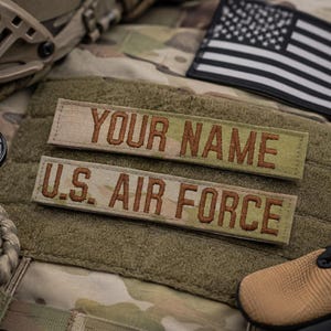 May include: Two rectangular tan fabric patches with brown embroidered text. The top patch reads YOUR NAME and the bottom patch reads U.S. AIR FORCE. These patches feature hook-and-loop backing and are positioned on a camouflage patterned surface.