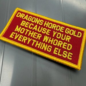 May include: A rectangular embroidered patch with a red background and a yellow border. The patch features yellow embroidered text that reads: DRAGONS HORDE GOLD BECAUSE YOUR MOTHER WHORED EVERYTHING ELSE. The design is stitched with a visible thread texture.