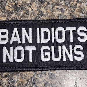 May include: A rectangular black patch with a stitched border featuring the white embroidered text BAN IDIOTS NOT GUNS arranged in two lines. The fabric has a visible grid-like texture, and the lettering is in a bold, sans-serif font style.