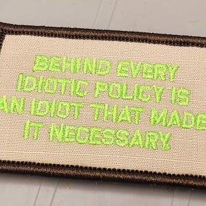 May include: A rectangular embroidered patch with a tan background and a dark brown stitched border. The patch features bright lime green embroidered text that reads: BEHIND EVERY IDIOTIC POLICY IS AN IDIOT THAT MADE IT NECESSARY in all capital letters.