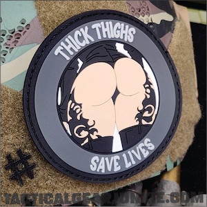 May include: A circular black tactical patch featuring a graphic of a person's lower back and thighs with black tribal-style tattoos. The text THICK THIGHS is printed in white at the top, and the text SAVE LIVES is printed in white at the bottom.