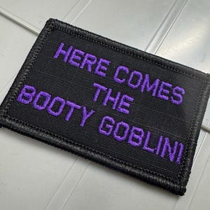 May include: A rectangular black embroidered patch with a thick black stitched border. The text "HERE COMES THE BOOTY GOBLIN!" is embroidered in a bold, purple sans-serif font across the center of the patch, creating a high-contrast design.