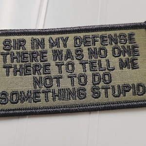 May include: A rectangular olive green embroidered patch with a black stitched border. The patch features black embroidered text that reads: SIR IN MY DEFENSE THERE WAS NO ONE THERE TO TELL ME NOT TO DO SOMETHING STUPID in a bold, sans-serif font.