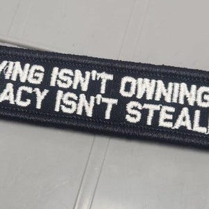 May include: A rectangular black embroidered patch with a stitched border. The text in white capital letters reads: IF BUYING ISN'T OWNING THEN PIRACY ISN'T STEALING. The patch features a dense fabric texture and clear, bold lettering throughout the design.