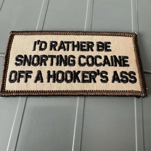 May include: A rectangular embroidered patch with a tan background and a dark brown border. The patch features black embroidered text that reads: I'D RATHER BE SNORTING COCAINE OFF A HOOKER'S ASS. The design is flat and has a visible fabric texture.