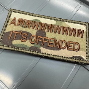 May include: A rectangular morale patch featuring a camouflage pattern in shades of green, brown, and tan. The patch has a dark olive green stitched border and displays the embroidered brown text AWWWWWWWWW IT'S OFFENDED in two lines.