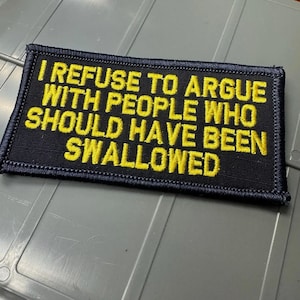 May include: A rectangular black embroidered patch with a dark border. The patch features bold yellow capital letters that read: I REFUSE TO ARGUE WITH PEOPLE WHO SHOULD HAVE BEEN SWALLOWED. The text is arranged in six lines across the center of the patch.