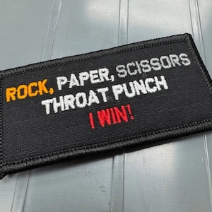 May include: A rectangular black embroidered patch with a stitched border. The text reads in orange, white, and red: ROCK, PAPER, SCISSORS THROAT PUNCH I WIN! The lettering is capitalized and centered on the fabric surface of the patch.