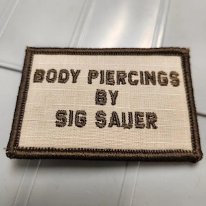 May include: A rectangular embroidered patch with a cream-colored background and a dark brown border. The patch features the dark brown embroidered text "BODY PIERCINGS BY SIG SAUER" arranged in three lines across the center of the fabric surface.
