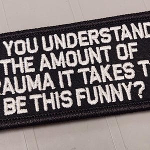 May include: A rectangular black embroidered patch with a stitched border. The patch features white embroidered text that reads: DO YOU UNDERSTAND THE AMOUNT OF TRAUMA IT TAKES TO BE THIS FUNNY? The lettering is in a bold, sans-serif font style.