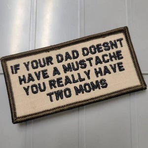 May include: A rectangular embroidered patch with a beige background and a dark brown border. The patch features black embroidered text that reads: IF YOUR DAD DOESNT HAVE A MUSTACHE YOU REALLY HAVE TWO MOMS. The lettering is in a bold, sans-serif font.