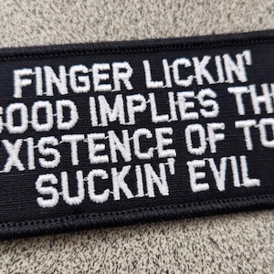 May include: A rectangular black embroidered patch with a stitched border. The patch features white text that reads: FINGER LICKIN' GOOD IMPLIES THE EXISTENCE OF TOE SUCKIN' EVIL. The lettering is in a bold, sans-serif font style across five lines.