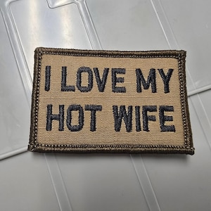 May include: A rectangular embroidered patch with a tan background and a dark brown border. The patch features the black embroidered text I LOVE MY HOT WIFE in a bold, sans-serif font. The design is centered on the fabric with a stitched edge finish.