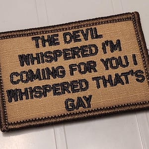 May include: A rectangular tan embroidered patch with a dark brown stitched border. The patch features black embroidered text that reads: THE DEVIL WHISPERED I'M COMING FOR YOU I WHISPERED THAT'S GAY. The design is centered on a textured fabric base.