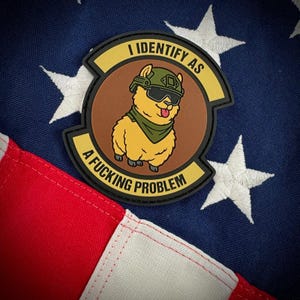 May include: A circular morale patch featuring a cartoon yellow llama wearing a green tactical helmet, black goggles, and a green neck scarf. The patch has a brown center and yellow borders with the black text I IDENTIFY AS A FUCKING PROBLEM.