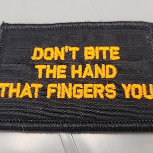 May include: A rectangular black embroidered patch with a stitched border. The text DON'T BITE THE HAND THAT FINGERS YOU is embroidered in bold yellow capital letters across the center of the patch, featuring a clear and legible font style.