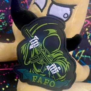 May include: A black and green tactical patch featuring a hooded figure holding a rifle with skeletal hands. The design includes a banner at the bottom with the text FAFO in yellow lettering. The patch has a dark border and a detailed graphic.