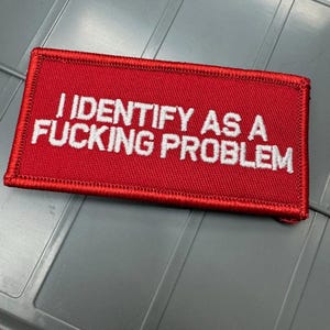 May include: A rectangular red embroidered patch with a stitched border. The patch features the white text "I IDENTIFY AS A FUCKING PROBLEM" in a bold, sans-serif font centered on the fabric surface, creating a high-contrast design.