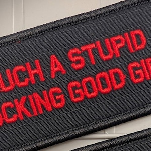 May include: A rectangular black embroidered patch with a stitched border. The text "SUCH A STUPID FUCKING GOOD GIRL" is embroidered in bold red capital letters across the center of the patch, featuring a textured fabric finish throughout.