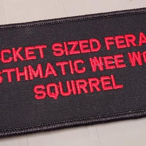 May include: A rectangular black patch with a stitched border featuring the red embroidered text POCKET SIZED FERAL ASTHMATIC WEE WOO SQUIRREL. The patch has a textured fabric surface and is designed for attachment to clothing or gear items.