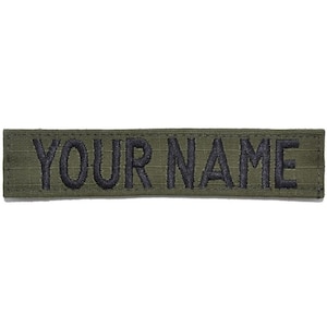 May include: A rectangular olive green fabric patch featuring the text YOUR NAME embroidered in bold black capital letters. The patch has a stitched border and a textured weave, designed for attachment to clothing or gear with a clean finish.
