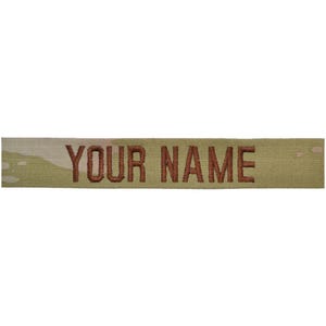 May include: A rectangular fabric name tape patch featuring a camouflage pattern in shades of tan, olive, and brown. The text "YOUR NAME" is embroidered in bold, dark brown capital letters across the center of the horizontal fabric strip.