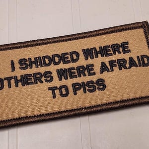May include: A rectangular tan morale patch with a dark brown embroidered border. The patch features black embroidered text that reads, I SHIDDED WHERE OTHERS WERE AFRAID TO PISS, arranged in three lines across the center of the fabric surface.