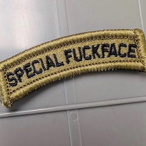 May include: A curved, olive drab embroidered patch featuring the text "SPECIAL FUCKFACE" stitched in bold, black, capitalized letters. The patch has a thick, stitched border in a matching olive green thread, creating a distinct, raised edge.