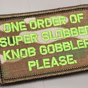 May include: A rectangular embroidered patch featuring a camouflage pattern in shades of brown, tan, and green. The patch has a dark brown stitched border and displays the text ONE ORDER OF SUPER SLOBBER KNOB GOBBLER, PLEASE in bright lime green.