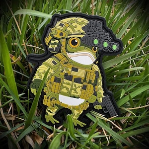 May include: A rubberized morale patch featuring a cartoon frog dressed in tactical gear, including a camouflage helmet with night vision goggles, a matching camouflage vest with a small American flag patch, and a black handgun held in one hand.